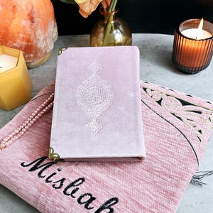 May include: A pink velvet prayer rug with the word "Misbah" embroidered in black. A matching pink velvet prayer book with gold accents and Arabic calligraphy is on top of the rug. A string of white pearls is on the rug.