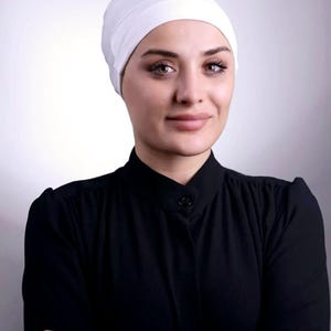 May include: A white head covering and a black long-sleeved shirt. The head covering is made of a soft, opaque fabric. The shirt has a button-down collar and a simple design. The person in the image has their arms crossed.