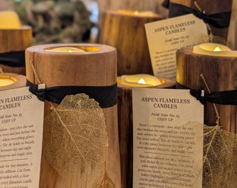 May include: Wooden candle holders with flameless candles. Each holder is cylindrical, made of light brown wood, and adorned with a black ribbon and a decorative leaf. The text "ASPEN FLAMELESS CANDLES" is visible on a tag.