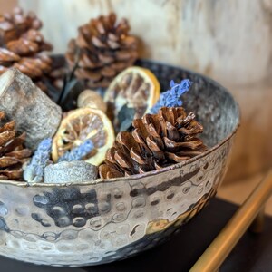 May include: A hammered metal bowl filled with pine cones, dried lemon slices, and sprigs of blue flowers. The bowl sits on a dark surface with gold-colored handles. The bowl's rim has a dark, aged appearance.