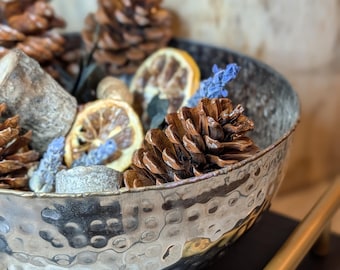 May include: A hammered metal bowl filled with pine cones, dried lemon slices, and sprigs of blue flowers. The bowl sits on a dark surface with gold-colored handles. The bowl's rim has a dark, aged appearance.