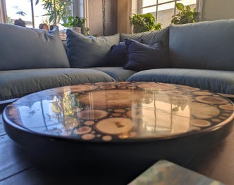 May include: A round coffee table with a glossy, clear surface. The table's top features a mosaic of wood slices encased in resin. The table has a dark, solid base and sits in front of a blue sectional sofa.