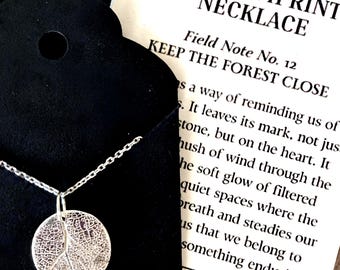 May include: A silver Aspen Imprint necklace with a circular pendant featuring a leaf design. The necklace is displayed on a black background with a card that reads "KEEP THE FOREST CLOSE". The necklace is made of sterling silver.