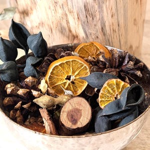 May include: A silver bowl filled with decorative items. The bowl contains dried orange slices, pine cones, dark blue leaves, and small wooden pieces. The arrangement is set against a wooden background.