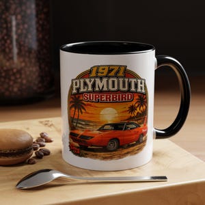 May include: A white ceramic mug with a black interior and handle, featuring a graphic of a 1971 Plymouth Superbird car parked on a beach at sunset. The text 1971 PLYMOUTH SUPERBIRD is printed in bold letters above the orange car and palm trees.