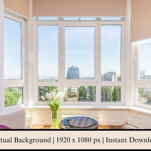 May include: A white living room with a large bay window overlooking a city skyline. The window has white blinds and white curtains. There are two white couches with pillows on them. There is a small table in front of the window with a vase of white flowers on it.