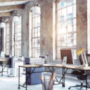 May include: An office interior with large windows, desks, and chairs. The room has a modern, industrial design with exposed beams and concrete walls. Desks have computers and lamps. The space is well-lit with natural light.