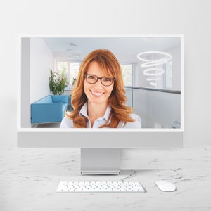 May include: A woman with red hair and glasses is on a video call on a computer monitor. She is wearing a white shirt and smiling. The background is a modern office with a blue sofa and a circular light fixture.