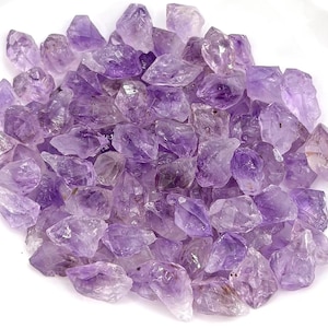 May include: A collection of raw amethyst crystal stones featuring various shades of translucent purple and violet. Each stone displays natural, jagged, and irregular geometric facets with varying degrees of clarity and depth of color throughout.