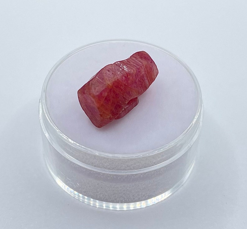 K&ouml;nnte beinhalten: Ein roher, facettierter Rubin-Edelstein, der einen tiefroten Farbton mit helleren Orange- und Rosat&ouml;nen aufweist. Der Rubin wird in einem transparenten, runden Kunststoffbeh&auml;lter mit wei&szlig;em Innenraum vor wei&szlig;em Hintergrund pr&auml;sentiert.