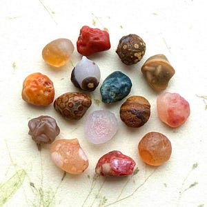 May include: A selection of polished gemstones in diverse shapes and colours. The stones exhibit shades of orange, red, brown, blue, and pink. Some have distinctive patterns and textures, creating a natural, earthy appearance.