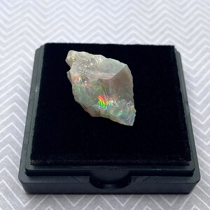 May include: A raw, iridescent opal crystal with a rainbow of colors, displayed on a black velvet background in a small, black box.