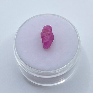 Peut inclure: Une pierre précieuse de rubis rose de forme brute et naturelle. Le rubis est présenté dans un petit récipient en plastique transparent.