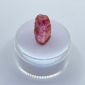 K&ouml;nnte beinhalten: Ein facettierter, r&ouml;tlich-rosa Kristall, pr&auml;sentiert in einem transparenten, runden Beh&auml;lter. Der Kristall hat eine nat&uuml;rliche, raue Textur mit Farbvariationen, die von tiefem Rosa bis zu helleren Farbt&ouml;nen reichen. Der Beh&auml;lter steht auf einer wei&szlig;en Oberfl&auml;che.