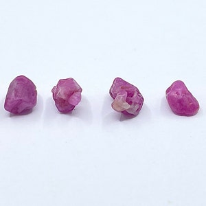 Puede incluir: Seis piedras preciosas en bruto de color rosa, probablemente rub&iacute;es, de diferentes tama&ntilde;os y formas, dispuestas en fila sobre una superficie blanca.