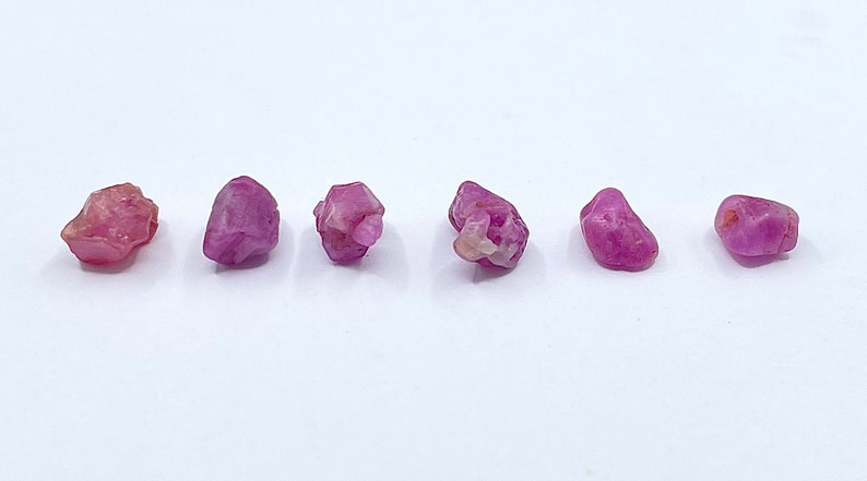 Puede incluir: Seis piedras preciosas en bruto de color rosa, probablemente rub&iacute;es, de diferentes tama&ntilde;os y formas, dispuestas en fila sobre una superficie blanca.