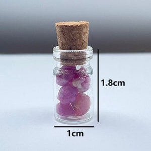 Puede incluir: Una pequeña botella de vidrio con un tapón de corcho llena de piedras preciosas rosas y rojas. La botella mide 1 cm de ancho y 1,8 cm de alto.