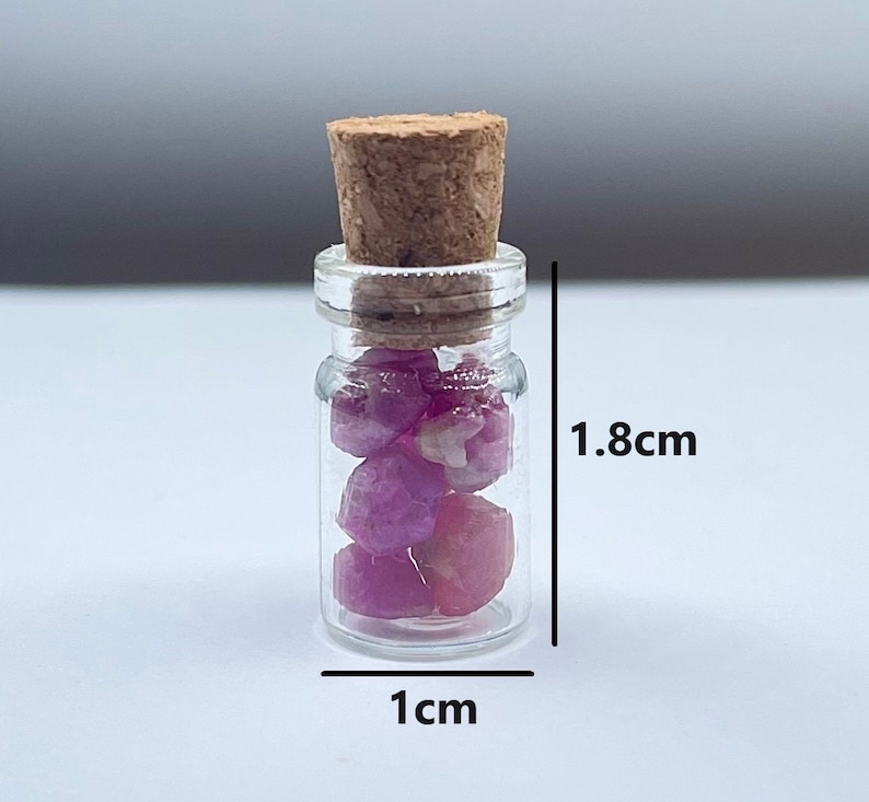 Puede incluir: Una peque&ntilde;a botella de vidrio con un tap&oacute;n de corcho llena de piedras preciosas rosas y rojas. La botella mide 1 cm de ancho y 1,8 cm de alto.
