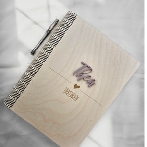 Könnte beinhalten: Ein Holzjournal mit einer hellen Holzoptik. Der Einband hat ein dekoratives Muster und das Wort "Thea" ist in Kursivschrift auf der Vorderseite geschrieben. Die Worte "Urkunden" sind ebenfalls auf der Vorderseite des Journals geschrieben.