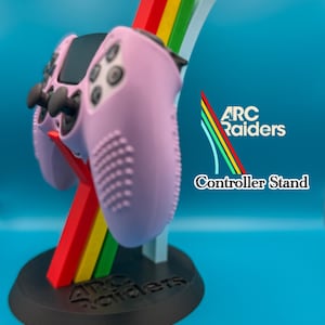 May include: A pink game controller rests on a black stand with the text "ARC Raiders" and "Controller Stand." The stand features a colorful, vertical design with red, yellow, green, and white stripes. The controller has black buttons and joysticks.