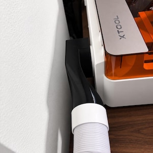 Puede incluir: Un adaptador de escape de plástico negro fijado al lateral de una máquina láser xTool blanca con tapa naranja transparente. Una manguera flexible blanca y corrugada está conectada al adaptador, extendiéndose hacia abajo desde el puerto lateral.
