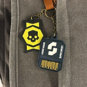 May include: Two keychains attached to a brown leather strap. The keychain on the left is a yellow and black star-shaped charm with a skull design. The keychain on the right is a blue and white rectangular charm with the text "SUPER CREDIT" and a stylized letter "S".