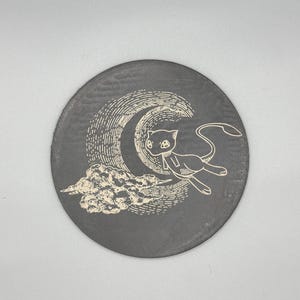 May include: A round, dark gray coaster featuring a white illustration of a cat-like creature, a crescent moon, and a cloud. The design is etched into the coaster's surface, creating a textured appearance. The coaster is a decorative item.