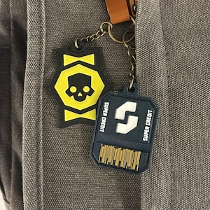 May include: A gray canvas bag featuring two hanging keychains. One is a yellow and black hexagonal charm with a skull icon. The other is a dark blue rectangular charm with a white logo, the text SUPER CREDIT, and a yellow barcode graphic.