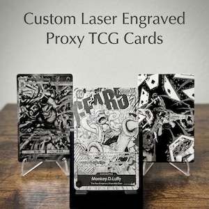 May include: Three custom laser engraved proxy TCG cards displayed on stands. The cards feature intricate black and white illustrations, including a central Monkey D. Luffy character card with the text 12000 and The Four Emperors/Straw Hat Crew.