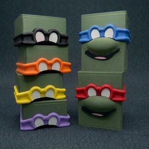 May include: A stack of four olive green rectangular blocks and two individual olive green blocks, each featuring a molded mask in black, orange, yellow, purple, blue, or red. The blocks with blue and red masks also display a molded nose and mouth.