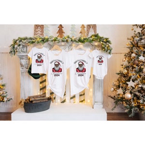 May include: Four white t-shirts featuring a black Santa Claus graphic and the text "Smith Family Christmas 2024". The shirts are displayed on a white shelf in front of a fireplace and Christmas trees.