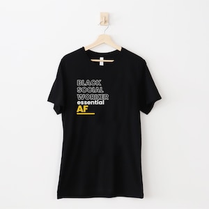May include: Black t-shirt with white text that reads "Black Social Worker Essential AF" in a bold, outlined font. The word "AF" is in yellow.