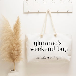 May include: White canvas tote bag with black text that reads "glamama's weekend bag" and "visit adore treat repeat".