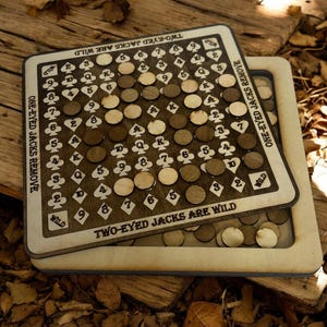 Könnte beinhalten: Ein Holzbrettspiel mit einer dunkelbraunen Spielfläche und hellbraunen Holzspielsteinen. Das Spielbrett trägt den Text "TWO-EYED JACKS ARE WILD" und "ONE-EYED JACKS REMOVE". Das Spiel befindet sich auf einer Holzoberfläche.
