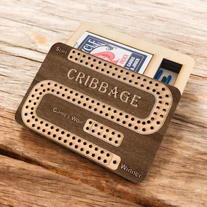 Op de afbeelding: Een houten cribbage-bordspel met een bruine afwerking. Het bord heeft een baan om punten te scoren, een "Start"-vak, een "Spellen gewonnen"-sectie en een "Winnaar"-sectie. Het bord is open om een ​​kaartspel en pionnen te onthullen.