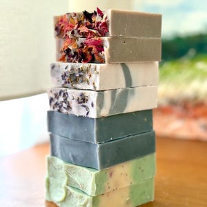 Puede incluir: Una pila de ocho barras de jabón artesanales en varios colores y patrones. La barra superior tiene pétalos de flores secas, las dos siguientes son grises, las dos siguientes son blancas con un patrón de espiral, las dos siguientes son azules y las dos inferiores son verdes.