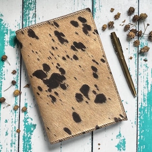 May include: A rectangular, tan leather journal with dark brown spots, resembling cowhide. A gold-colored pen rests beside the journal. The journal is closed and has a stitched border. The background is a distressed, painted wooden surface.
