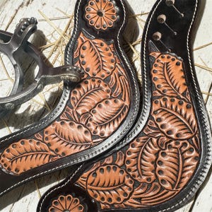 May include: Close-up of two brown leather spur straps with intricate leaf and floral tooling. The straps have a black border with white stitching and are paired with a silver spur.