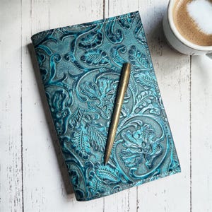 May include: A teal leather journal with an embossed floral design and a gold pen resting on top. The journal is open, revealing blank pages. A cup of coffee sits in the background.