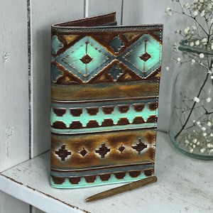 May include: A handcrafted leather journal with a turquoise and brown geometric pattern. The journal is open, revealing blank pages, and a pen rests beside it. The cover features a detailed, embossed design, showcasing a rustic, Southwestern aesthetic.