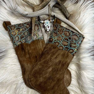 May include: Two brown cowhide Christmas stockings with turquoise and gold accents. The stockings are decorated with a faux alligator skin pattern. The stockings are displayed on a white faux fur background with two antler ornaments.