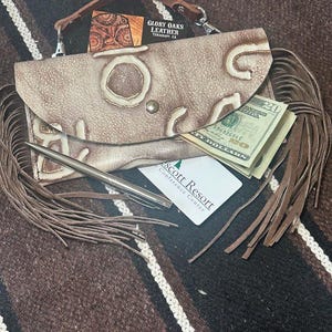 May include: A tan leather clutch with a handle, fringe, and decorative cutouts. The clutch contains a pen, cash, and a card. The text "GLORY OAKS LEATHER TERRACH CA" is visible on a small card.