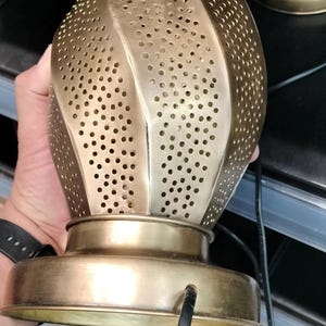 May include: A gold-colored metal lantern with a perforated design. The lantern has a bulbous shape with a flat base and a black electrical cord. The design features small, evenly spaced holes throughout the body.