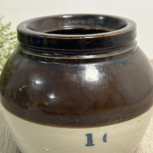 Puede incluir: Un tarro de gres vintage marrón y crema. La parte superior es marrón brillante, la mitad inferior crema con un "1" azul impreso en el lateral. Tiene una amplia abertura y una forma redondeada. Ideal para la cocina.