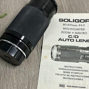 May include: A black Soligor 80-200mm/F4.5 multicoated zoom + macro C/D auto lens with a textured grip. The lens is next to a manual with the same lens pictured and text in multiple languages.
