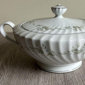 Puede incluir: Tazón de servir de porcelana blanca con tapa, con un delicado patrón floral en tonos verdes y azules. El tazón tiene un diseño acanalado y un borde dorado.