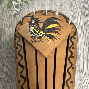 May include: Wooden rooster-themed kitchen utensil holder. The holder is made of vertical wooden slats with a decorative black wavy pattern along the edges. A painted rooster is on the top, featuring black, yellow, and white details. The holder is approximately 7 inches tall.