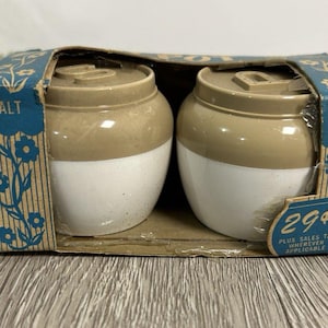 May include: A vintage salt and pepper shaker set in a cardboard box. The ceramic shakers are two-toned, with a white base and tan top. The box is blue with floral designs and the words "SALT" and "PEPPER".