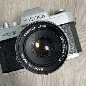 May include: A silver and black Yashica FX-2 camera with a black lens. The lens is marked "Yashica Lens" and "DSB 50mm 1:1.9". The camera is labeled "Made in Japan".