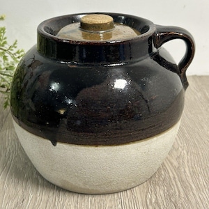 Puede incluir: Un tarro de cerámica vintage con tapa. El tarro es de dos tonos, con una sección superior marrón oscuro y una sección inferior de color crema. Tiene un asa y un tapón de corcho. El tarro es probablemente para almacenamiento o decoración.